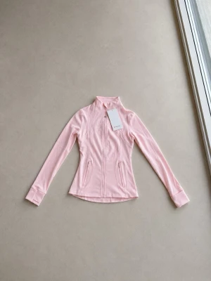 Ljusrosa zip-jacka från Lululemon - Supersnygg ljusrosa träningsjacka från Lululemon med figurnära passform, hel dragkedja framtill och två fickor med dragkedja. Tillverkad i ett stretchigt och mjukt funktionsmaterial som passar perfekt till träning eller som ett extra lager.