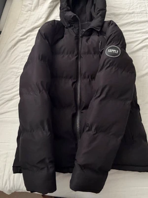 Svart pufferjacka Supply & Demand - Säljer en svart pufferjacka med huva och dragkedja framtill. Jackan har två sidofickor och en patch med texten 'SUPPLY' på ena ärmen. Perfekt för kalla dagar och har en klassisk, oversized look.