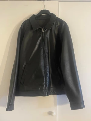 Svart klassisk skinnjacka - Snygg svart skinnjacka med rak passform och dragkedja framtill. Jackan har två snedställda fickor och en stilren krage. Perfekt för dig som vill ha en tidlös och enkel look med edge. Är en ”mans jacka” men jag som tjej använde den som baggy skinnjacka bara 