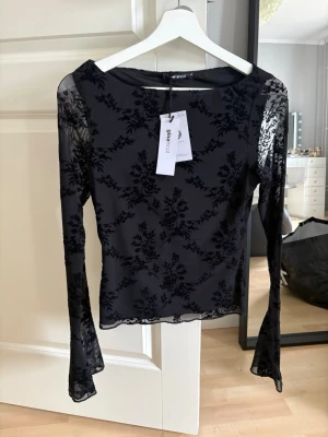 Svart meshblus med blommigt mönster - Snygg svart meshblus med transparenta långärmar och blommigt sammetstryck. Blusen har en figurnära passform och vida ärmslut för en trendig look. Perfekt att styla med jeans eller kjol för en cool outfit.