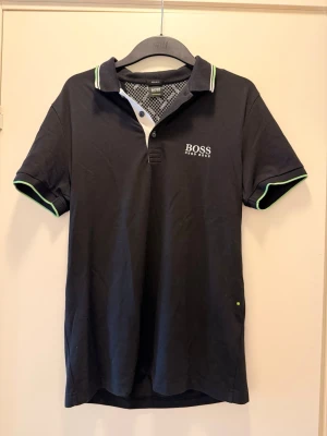 Svart pikétröja från Hugo Boss M - Snygg svart pikétröja från Hugo Boss med vit logga på bröstet. Tröjan har krage med vita och gröna detaljer samt korta ärmar med gröna kanter. Tillverkad i mjuk bomull, perfekt för en clean och sportig look.