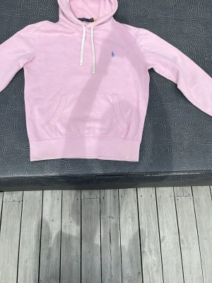 Ljusrosa hoodie från Polo Ralph Lauren - Snygg ljusrosa hoodie från Polo Ralph Lauren med klassisk blå logga broderad på bröstet. Hoodien har vit dragsko, känguruficka och mjukt material som känns skönt mot huden. Perfekt för skolstarten. 