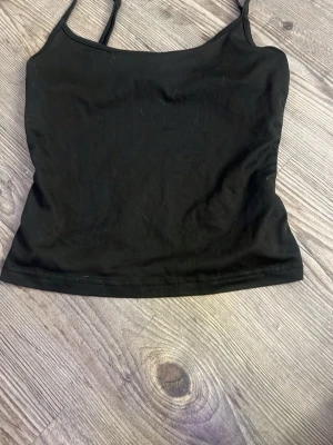 Svart ärmlös linnetopp - Säljer en enkel och stilren svart linnetopp med smala axelband. Toppen har en tight passform och är perfekt att matcha med jeans eller kjol. Materialet känns mjukt och stretchigt, vilket gör den bekväm att bära hela dagen.