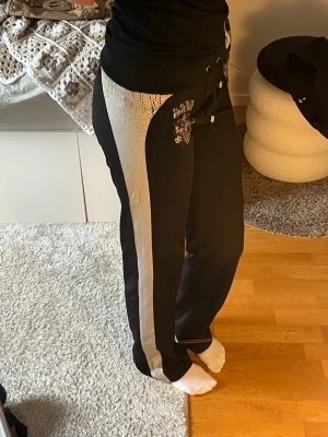 Svarta mjukisbyxor med beige detalj - Säljer ett par svarta mjukisbyxor med bred beige sidopanel och coolt tryck med texten 'BERG' och blingade detaljer på ena benet. Byxorna har dragsko i midjan och avslappnad passform. Perfekta för en chill och trendig look.