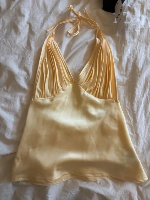 Ljusgul halterneck linnetopp i satin - Säljer en ljusgul linnetopp i satin med halterneck och djup v-ringning. Toppen har tunna knytband i nacken och ett glansigt, slätt tyg som ger en lyxig känsla. Perfekt för dig som vill sticka ut med en elegant och trendig topp.