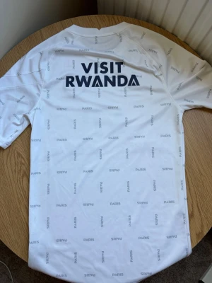 Vit PSG fotbollströja Jordan - Säljer en vit PSG fotbollströja i sportigt material med tryck i svart och grått. Tröjan har Paris Saint-Germain-logga, Jordan-logga och texten 'Accor Live Limitless' på framsidan samt 'Visit Rwanda' på ryggen. Perfekt under sommarn och för dig som älskar fotboll och vill ha en snygg supportertröja. 
