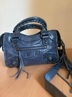 Blå handväska i läder med nitar - Cool blå handväska i läder med flätade handtag, dekorativa nitar och dragkedja framtill. Väskan har både axelrem och handtag samt långa läderdetaljer som ger en edgy look. Perfekt för dig som vill sticka ut med en unik accessoar.