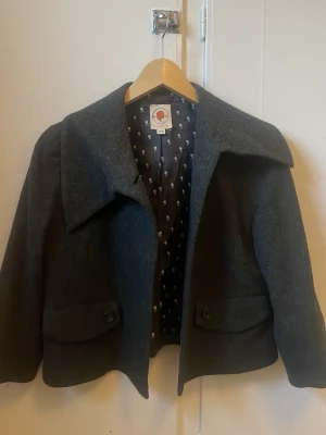 Svart supergullig poncho - Snygg mörkgrå kappa i ullblandning med stor, asymmetrisk krage och två stora fickor framtill med knappar. Fodret har ett diskret mönster. Perfekt för kyliga dagar och ger en stilren look.