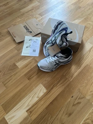 Asics  - Snygga vita Asics sneakers med svarta lila och silvriga detaljer. Skorna har en sportig design med mesh- och syntetmaterial för bra andningsförmåga. Perfekta för dig som gillar en clean och modern look. Levereras med originalkartong och tags.