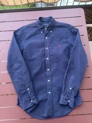 Polo Ralph Lauren skjorta  - Riktigt snygg Polo Ralph Lauren skjorta i storleken S. Inga tydliga defekter som jag har märkt. Fin marinblå färg med röd polo logga på bröstet. Riktigt skön och fungerar perfekt nu under sommaren. Pris går att diskutera vid snabb affär. Skickas inom 24h.