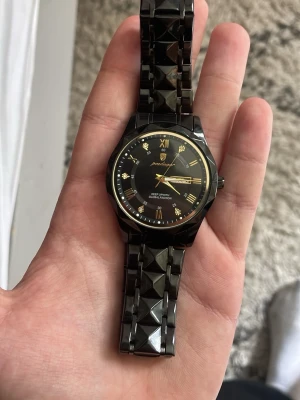 Svart klocka med gulddetaljer - Stilren svart klocka med guldfärgade romerska siffror och visare. Armbandet har en unik geometrisk design som ger en cool och modern känsla. Perfekt accessoar för dig som vill sticka ut lite extra.