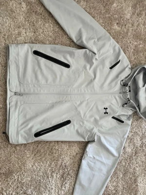 Under armour S/FITCH - Snygg ljusgrå vindjacka från S/FITCH med svart dragkedja och detaljer. Jackan har huva med dragsko, två snedställda fickor framtill och meshfoder på insidan. Perfekt för blåsiga dagar och enkel att matcha med olika outfits.