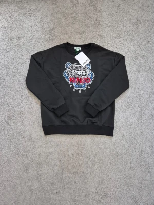 Kenzo - Cool svart sweatshirt från Kenzo med det ikoniska tigerhuvudet broderat framtill i vitt, blått och rött. Rund halsringning och ribbade muddar vid ärmslut och nederkant. Perfekt för en trendig streetwear-look.