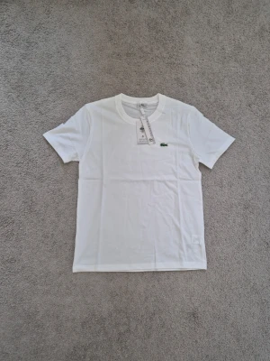 Lacoste - Klassisk vit t-shirt från Lacoste med rund halsringning och det ikoniska gröna krokodilmärket broderat på bröstet. Tillverkad i mjuk bomull och har en enkel, stilren design med korta ärmar. Perfekt för en clean och avslappnad look.