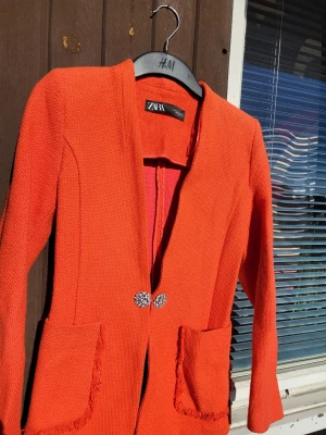 Orange kavaj från ZARA med smyckesknappar - Snygg orange kavaj från ZARA i storlek XS. Kavajen har två fickor framtill med fransade kanter och stängs med två dekorativa smyckesknappar. 