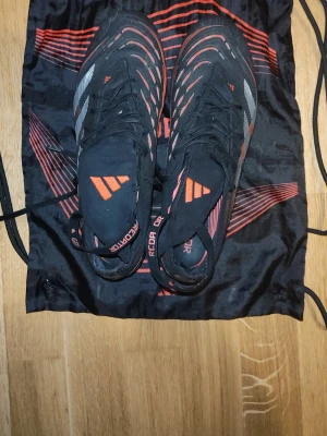 Adidas Predator ft elite 25 fotbollsskor - Säljer ett par svarta Adidas Predator fotbollsskor med röda och vita detaljer. Skorna har snörning och en modern design med mönstrade linjer över ovandelen. Perfekta för dig som vill ha stil och kontroll på planen. 