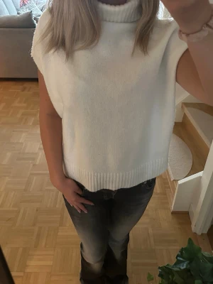 Vit stickad ärmlös polotröja - Säljer en vit stickad ärmlös polotröja med hög krage. Tröjan har en avslappnad passform och ribbade detaljer vid krage och nederkant. Perfekt att styla över en långärmad topp eller t-shirt för en trendig lager-på-lager-look.