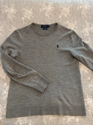 Grå stickad tröja Polo Ralph Lauren Merinoull - Snygg grå stickad tröja från Polo Ralph Lauren med rund halsringning och  broderad logga på bröstet. Tröjan har långa ärmar och ribbade muddar vid ärmslut och nederkant. Perfekt basplagg i garderoben. OBS liten o storleken, S-M. 94% merinoull.