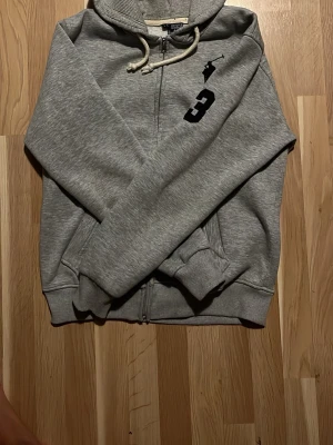 Grå hoodie från Polo Ralph Lauren (sällsynt) - Säljer en grå hoodie från Polo Ralph Lauren med dragkedja, huva och vita snören. På bröstet finns en svart logga och siffran 3. Hoodien har ribbade muddar och är gjord i mjukt material, perfekt för en chill och avslappnad stil. Storlek L fast passar som M