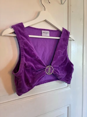 Lila sammetstopp från Juicy Couture - Säljer en lila croppad sammetstopp från Juicy Couture med breda axelband och en snygg ringdetalj framtill. Toppen har en djup v-ringning och är perfekt för dig som vill sticka ut med en färgstark och mjuk look. Den är liten i storleken och passar snarare en S/M.