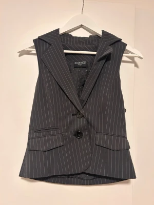 Svart randig kostymväst från Soaked - Snygg svart kostymväst/blazer med gråa ränder från Soaked. Klassisk kavajkrage, två knappar framtill och fickdetaljer. Perfekt för att lyfta din outfit med en stilren och trendig touch. Snygg figursydd passform!
