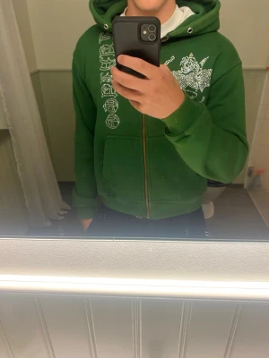 Grön hoodie med dragkedja från Laviago - Grön hoodie från Laviago med dragkedja framtill och vit tryckt design på bröstet och längs ena sidan. Klassisk huva och kängurufickor. Perfekt för en chill och avslappnad stil.
