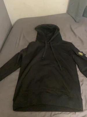 Svart hoodie med Stone Island patch - Säljer en svart hoodie från Stone Island med klassisk patch på vänster ärm. Hoodien har dragsko i huvan och ribbade muddar. Perfekt för dig som gillar streetwear och vill ha en stilren look. Materialet känns mjukt och bekvämt.