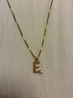Guldhalsband med bokstav E och sten - Snyggt halsband i guldton med ett hänge i form av bokstaven E. Hänget har en liten klar sten i nedre hörnet som ger extra bling. Kedjan är tunn och elegant, och hänget är stämplat med 925 vilket tyder på att det är i silver med guldfinish.