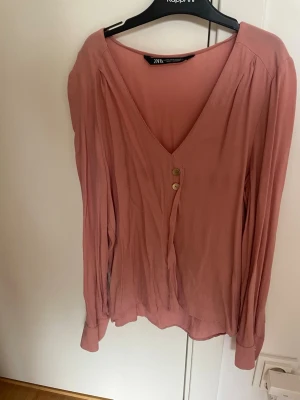 Rosa blus med knappar från Zara - Snygg rosa blus från Zara med djup V-ringning och två dekorativa knappar framtill. Blusen har långa ärmar och är tillverkad i ett lätt, mjukt tyg som faller fint. Perfekt att styla med jeans eller kjol för en trendig look.