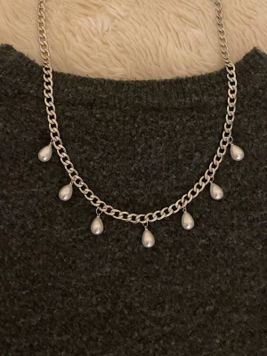 Silverfärgat halsband med droppformade pärlor - Stilrent halsband i silverfärgad rostfritt med kedja och sju droppformade vita pärlor som hänger längs kedjan. Perfekt accessoar för att lyfta din outfit med en elegant touch.