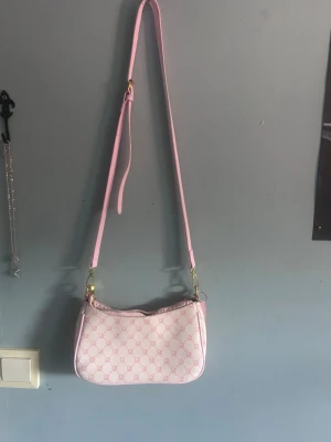 Ljusrosa axelremsväska med mönster - Söt ljusrosa handväska med justerbar axelrem och guldfärgade detaljer. Väskan har ett diskret mönster i rosa och vitt samt dragkedja upptill. Perfekt för dig som gillar en feminin och trendig look.