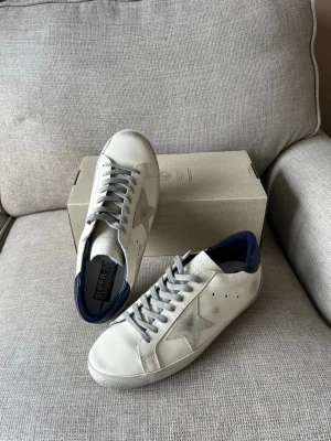 Vita sneakers med stjärndetalj - Snygga vita sneakers med grå stjärndetalj på sidan och mörkblå häl. Skorna har grå skosnören och perforerade detaljer för extra stil. Tillverkade i läder med textilfoder och låg profil. Perfekta för dig som gillar en clean och trendig look.
