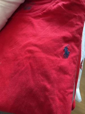 Röd Ralph lauren t shirt - En klassisk röd t-shirt med rund hals och korta ärmar. På bröstet finns en liten broderad logga i blått som ger en stilren touch. Tillverkad i mjuk bomull för skön känsla hela dagen. Perfekt till jeans eller shorts. Står storlek S men skulle säga den passar xs då den är liten i storlek