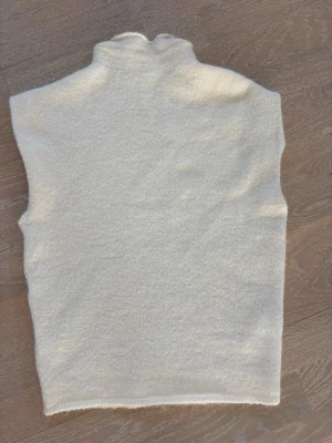 Vit ullväst från Gina Tricot - Mysig vit väst från Gina Tricot i ullblandning. Västen har hög krage och är enkel i modellen utan knappar eller dragkedja. Perfekt att styla över en skjorta eller tröja för en clean och trendig look.