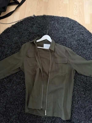 Olivgrön overshirt från Ciszere - Stilren olivgrön overshirt från Ciszere med dragkedja framtill och två bröstfickor med lock. Skjortkrage och långa ärmar med knapp vid ärmslut. Tillverkad i ett tunt, mjukt tyg som ger en avslappnad look. Ny pris 1 099 Storlek L / M funkar 