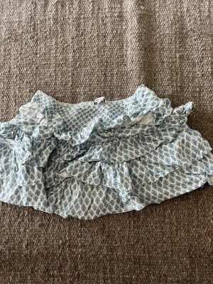 Blåvit volangkjol från Subdued - Supersöt kort kjol från Subdued i blått och vitt med småblommigt mönster och flera volanger. Kjolen har smock i midjan för skön passform och är perfekt för varma dagar. Tillverkad i ett lätt och luftigt material.