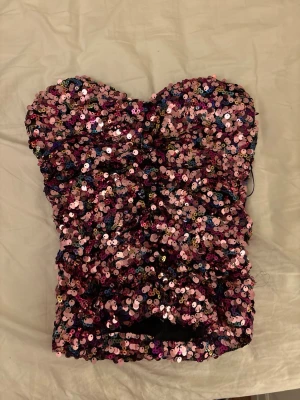 Glittrig paljett tubtopp i rosa - Säljer en superglittrig tubtopp täckt med rosa, lila och silvriga paljetter. Toppen har en bandeauform och tunna axelband som kan användas eller stoppas in för axelbandslös look. Perfekt för dig som vill sticka ut!