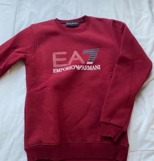 Vinröd sweatshirt från Emporio Armani - Snygg vinröd sweatshirt från Emporio Armani EA7 med tryck på bröstet. Tröjan har rund halsringning, ribbade muddar och är tillverkad i mjukt material som känns skönt mot huden. Perfekt för en avslappnad och trendig look.