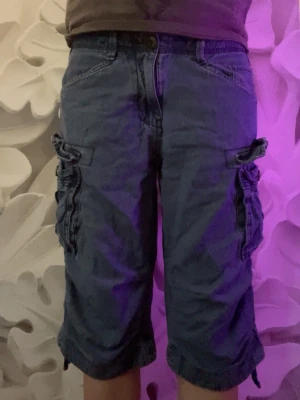 Blå cargoshorts i jeans - Säljer ett par blå cargoshorts i jeansmaterial med flera stora fickor på sidorna och bak. Shortsens längd går strax under knäna och de har en klassisk knapp och dragkedja framtill. Perfekta för en avslappnad stil.