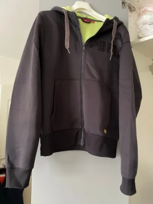 Svart hoodie med dragkedja från Puma - Snygg svart hoodie från Puma med dragkedja framtill, fickor och huva med snören. Insidan av huvan är limegrön för en cool kontrast. Puma-logga i limegrönt på ryggen. Perfekt för en avslappnad och sportig stil. Polyester.