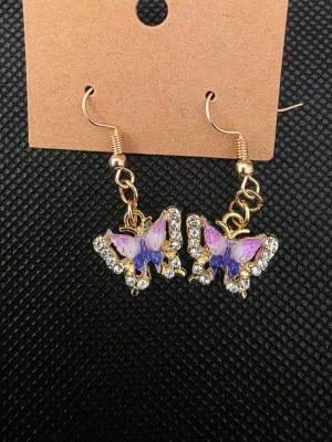 Lila fjärilsörhängen med strass - Snygga örhängen med fjärilar i lila och rosa nyanser, dekorerade med glittrande strass. Hängande modell med guldfärgade detaljer och krok. Perfekta för att ge din look en färgklick och lite extra bling.