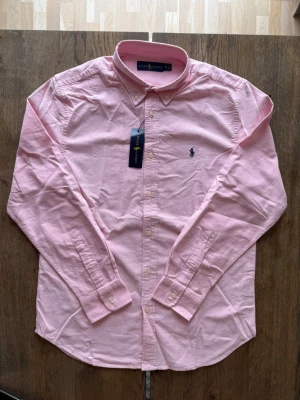 Rosa skjorta från Ralph Lauren - Klassisk rosa skjorta från Ralph Lauren med button-down krage och broderad logga på bröstet. Skjortan har långa ärmar, knappar framtill och är tillverkad i mjuk bomull. Perfekt för en stilren och avslappnad look.