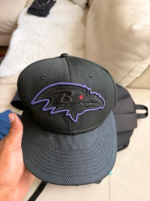 Svart Baltimore Ravens keps - Snygg svart keps från New Era med Baltimore Ravens-logga broderad framtill i lila och svart. Kepsen har böjd skärm och är tillverkad i ett ventilerande mesh-liknande material. Perfekt för dig som gillar amerikansk fotboll och vill ha en sportig look.