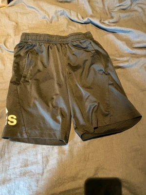 Svarta träningsshorts från Adidas - Snygga svarta träningsshorts från Adidas med elastisk midja och två sidofickor. Stora vita Adidas-loggor på ena benet. Tillverkade i ett lätt och snabbtorkande material som passar perfekt för sport och aktivitet.