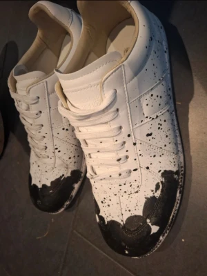 Maison margielas med svart färgstänk - Unika vita sneakers med svart färgstänk och målade detaljer på tån. Skorna har klassisk låg profil, vita snören och ovandel i slätt material. Perfekta för dig som vill sticka ut med en kreativ och edgy stil.