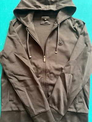 Svart hoodie med dragkedja Emporio Armani - Svart hoodie från Emporio Armani med dragkedja framtill, justerbar huva med snören och diskret logomärke på bröstet. Tillverkad i mjukt bomullsmaterial och har två fickor på sidorna. Perfekt för en avslappnad och stilren look.