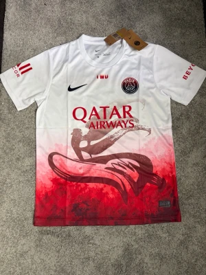 PSGfotbollströja  - Säljer en snygg tröja i vitt och rött. Tröjan har korta ärmar, , Qatar Airways-tryck fram och Visit Rwanda-tryck bak. Unik grafisk design i rött nedtill och detaljer på ärmarna. Perfekt för fotbollsfans!