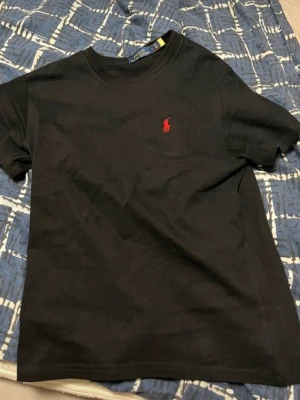 Polo Ralph lauren t shirt  - Svart t-shirt i bomull med klassisk rund hals och korta ärmar. På bröstet finns en liten röd broderad logga som ger en stilren touch. Perfekt för en enkel och snygg look.