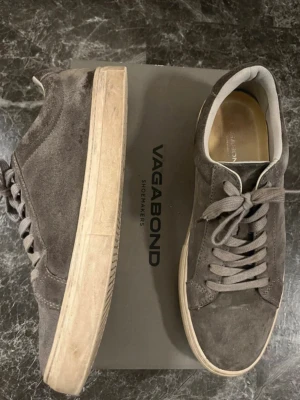 Bruna Vagabond sneakers i mocka - Snygga bruna sneakers från Vagabond i mjuk mocka med klassisk låg profil och beige sula. Skorna har matchande bruna skosnören och diskreta sömmar för en clean look. Perfekta för dig som gillar stilrena och tidlösa sneakers.