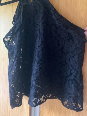 Svart spetsig ärmlös topp - Snygg svart topp i spets med hög hals och ärmlös design. Blommigt spetsmönster ger en elegant vibe och toppen har en lätt transparent look. Perfekt att styla med jeans eller kjol för en fest  outfit.